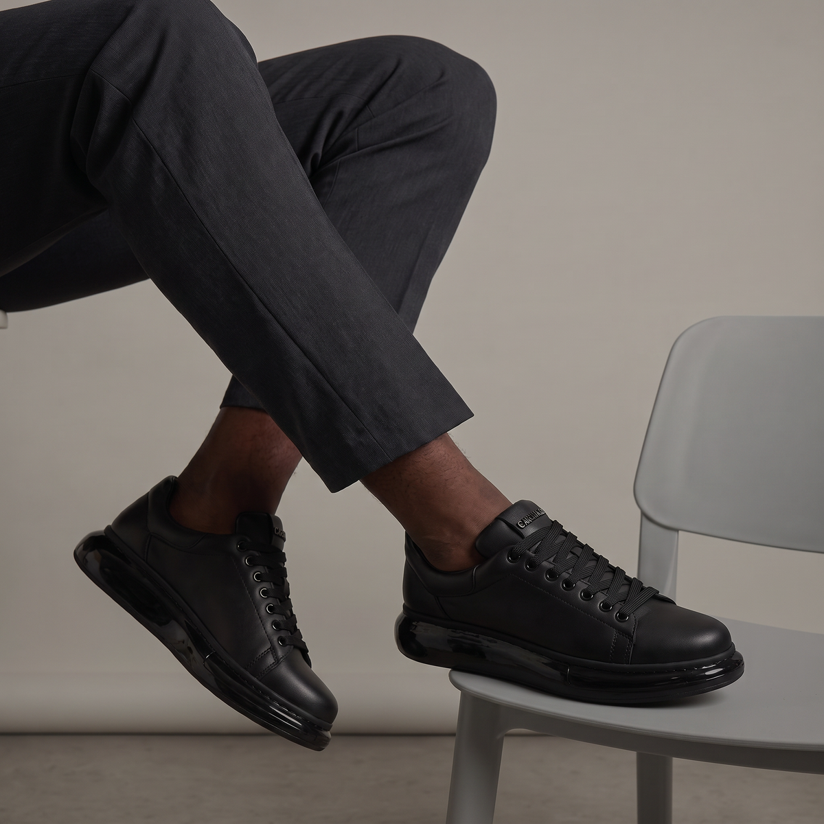 Chekich FANTASIA X All Black sneakers worn with grey trousers, seated on a chair